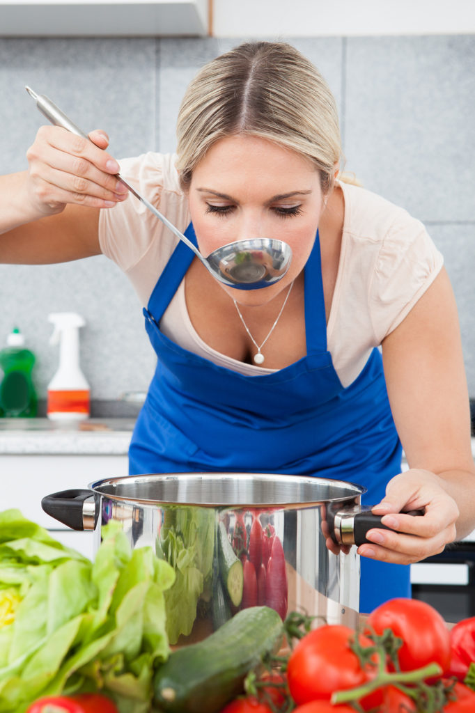 woman leaning over and tasing broth from a stainless steel stock pot with veggies in the foreground